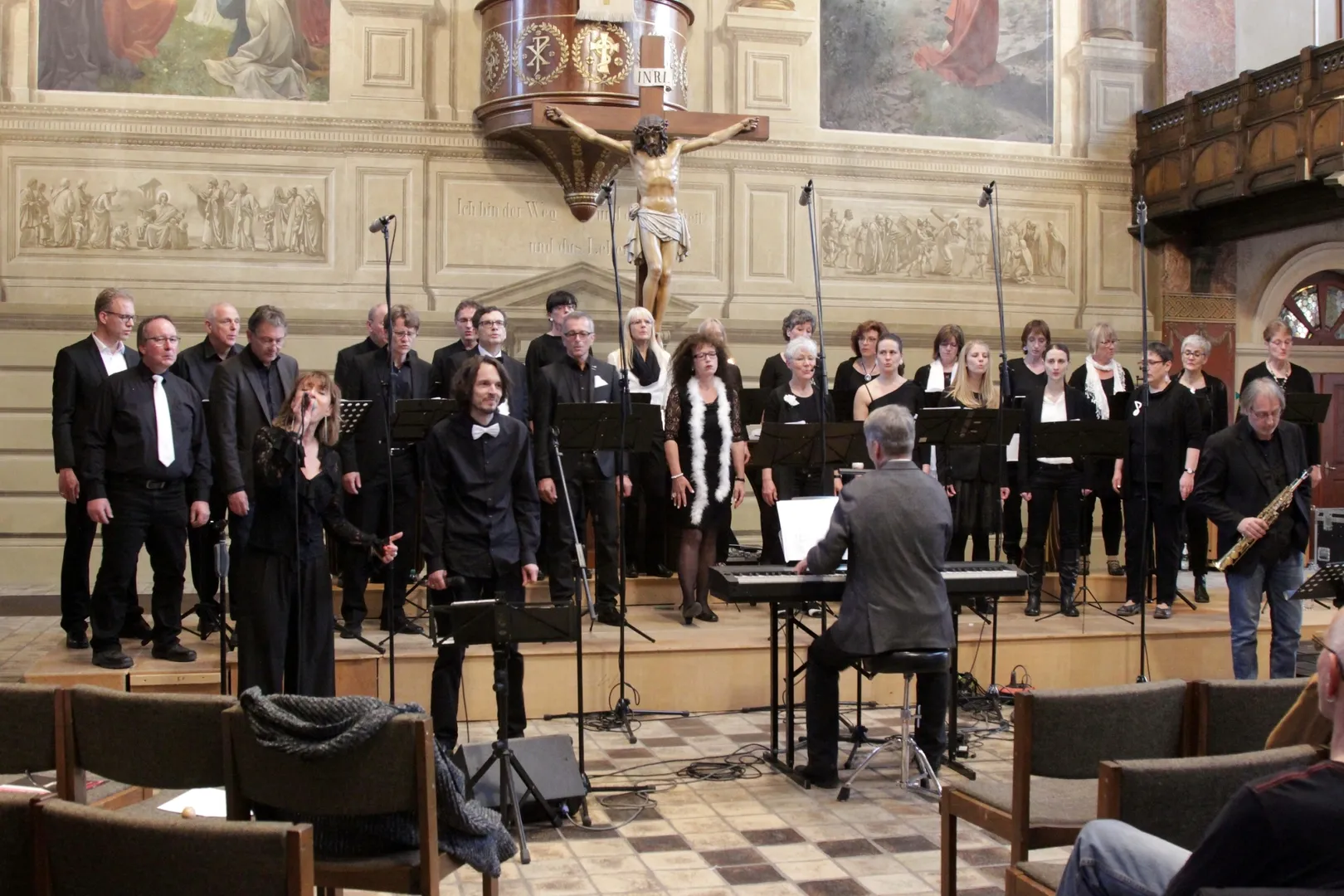 Konzert in der Stadtkirche Tuttlingen: der Chor in Schwarz vor Fresken und Kruzifix, vorne eine Solistin am Mikrofon und ein Sänger, am Keyboard der Chorleiter von hinten, rechts ein Saxophonist.